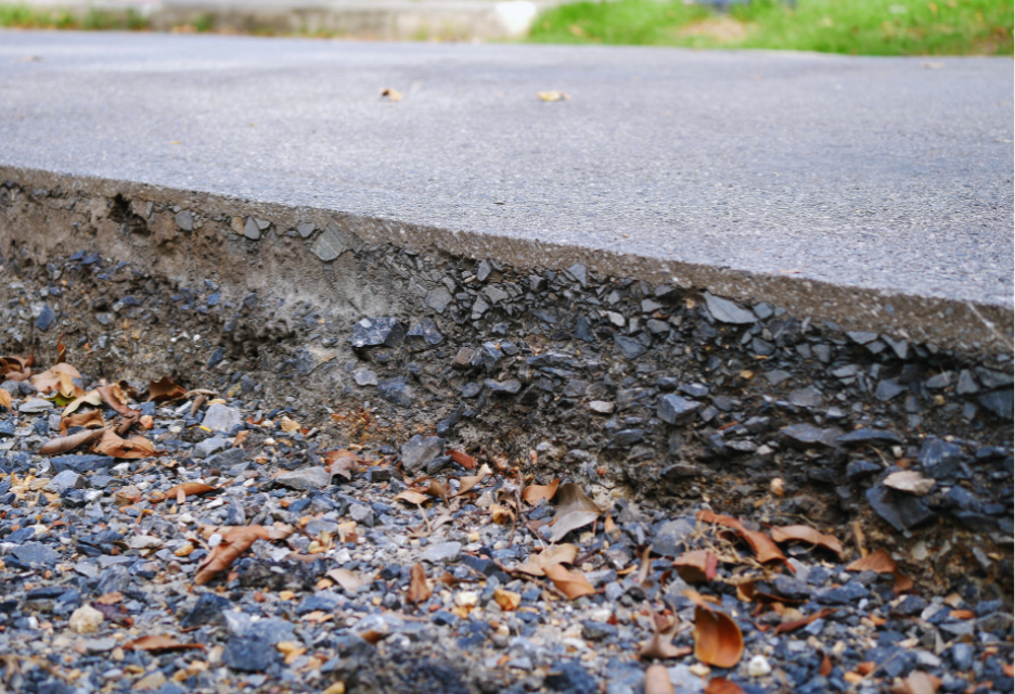 Close-up cross-section of an asphalt road showing the pavement layer and compacted aggregate base beneath it, with gravel and fallen leaves along the exposed edge.